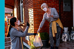 Two women in masks where one is handing the other a bag of groceries